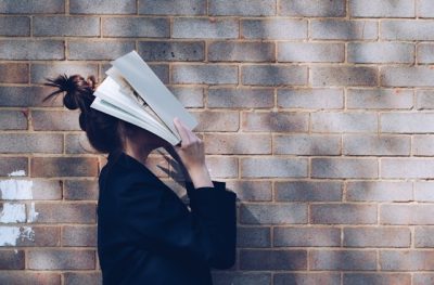 Junge Frau hält sich ein aufgeschlagenes Buch vor das Gesicht und steht vor einer Backsteinwand.