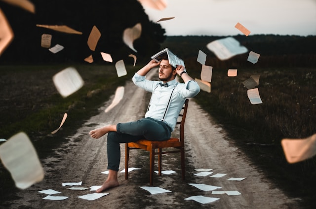 Student mit Buch auf dem Kopf, umgeben von fliegenden Blättern – sinnbildlich für Prüfungsstress und Vorbereitungschaos.